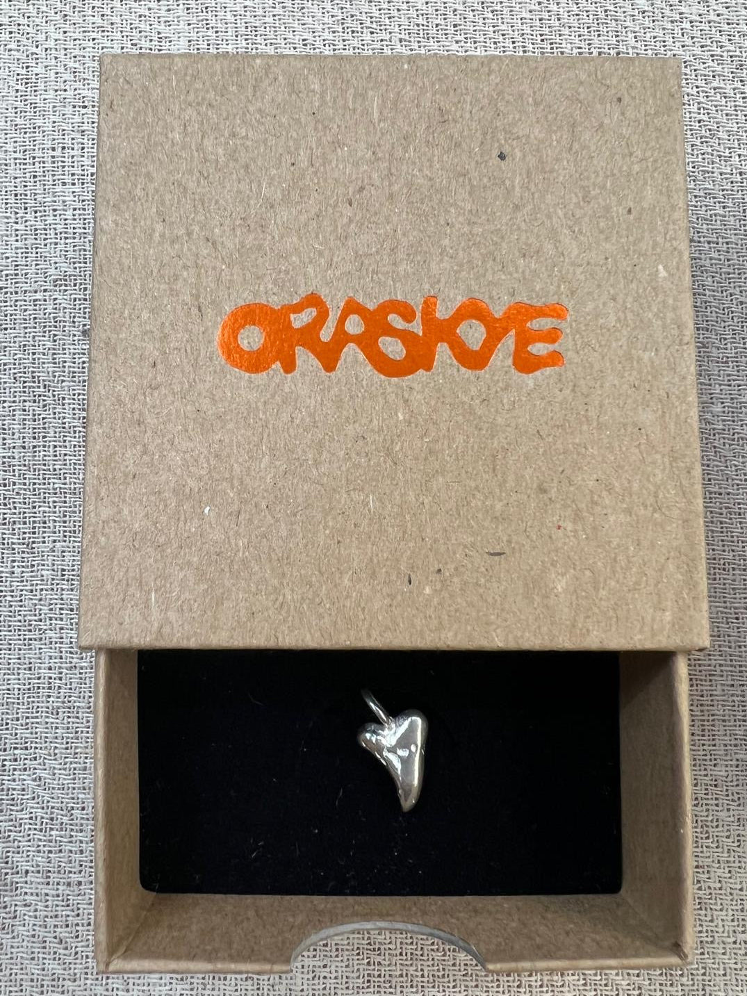 Silver dog tooth charm in a cardboard box with 'Oraskye' branding.