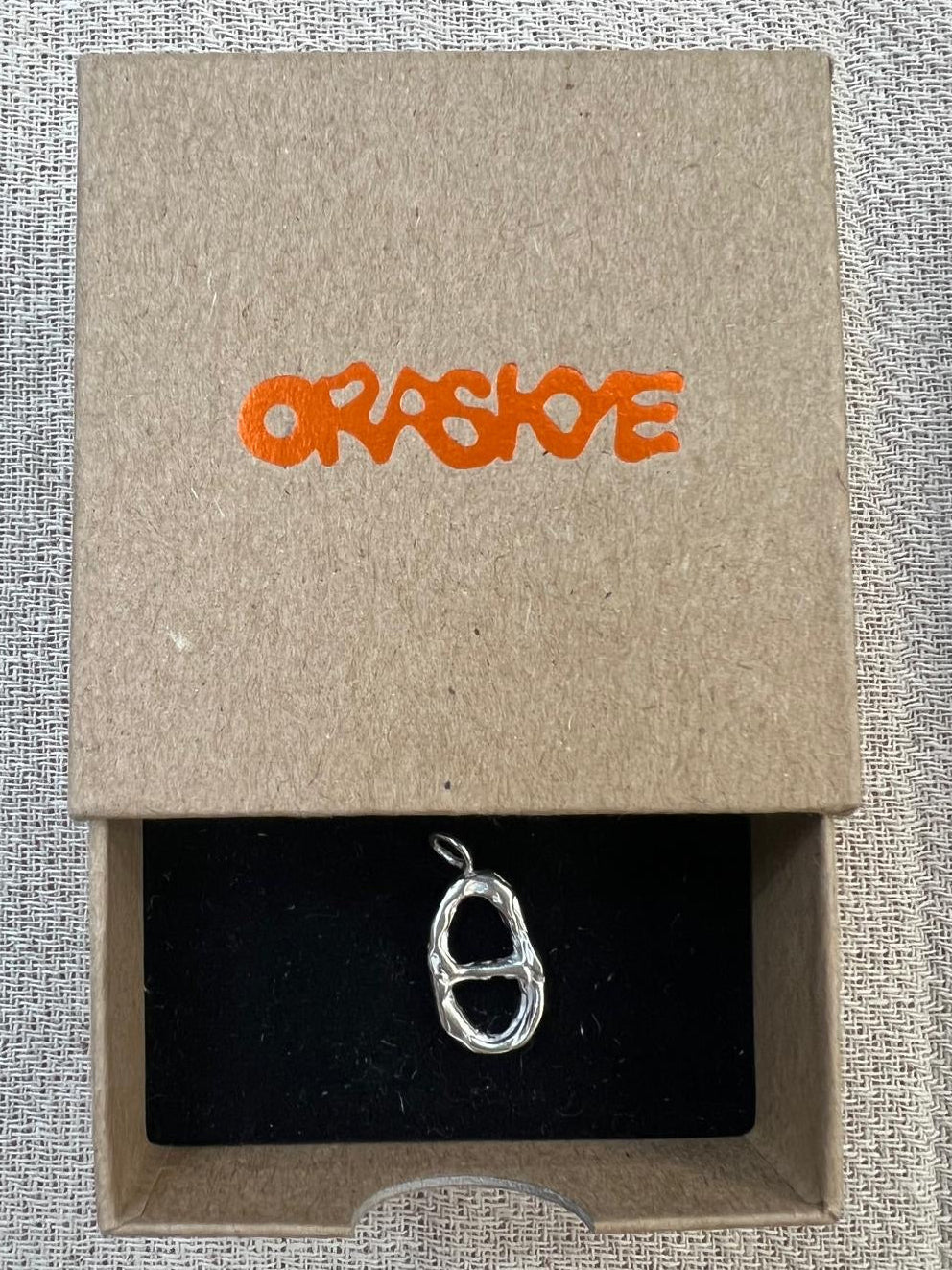 Silver tab charm in a 'ORASKYE' branded box on a textured gray background