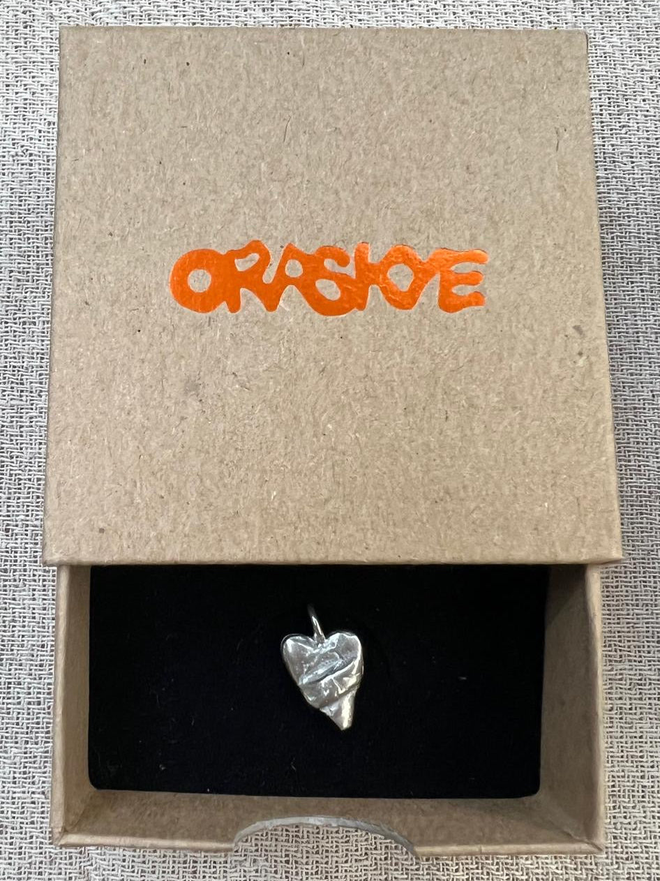 Silver heart-shaped pendant in a cardboard box with 'ORASKYE' branding.