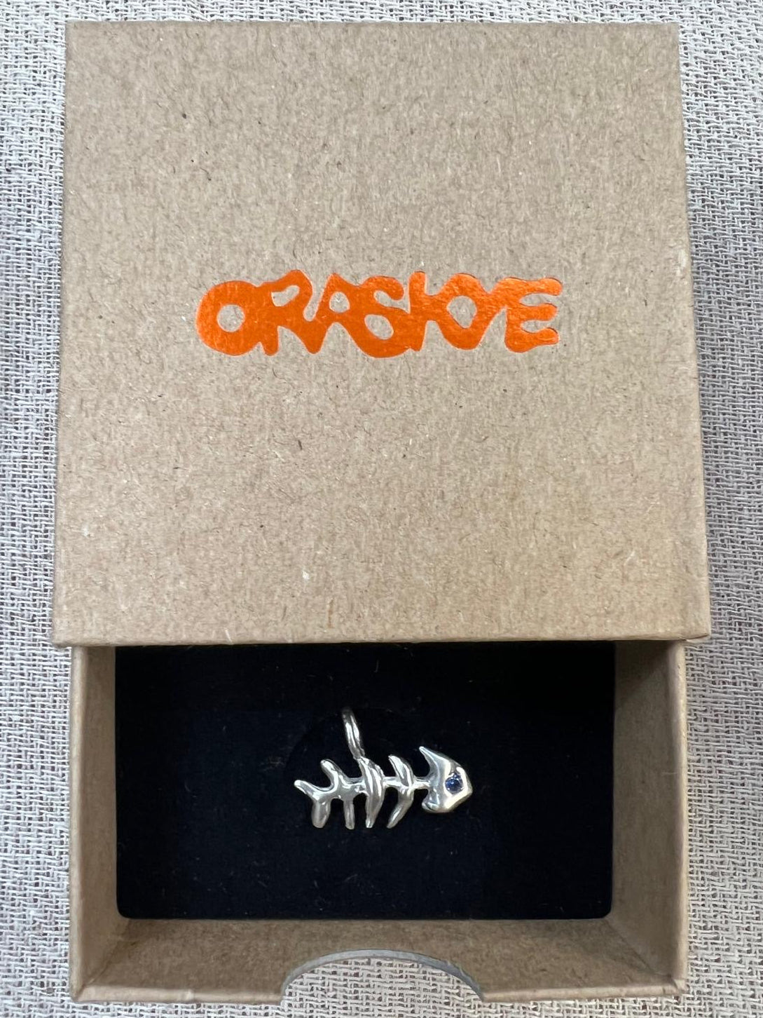 Silver fish bone charm in a 'ORASKYE' branded box on a textured surface