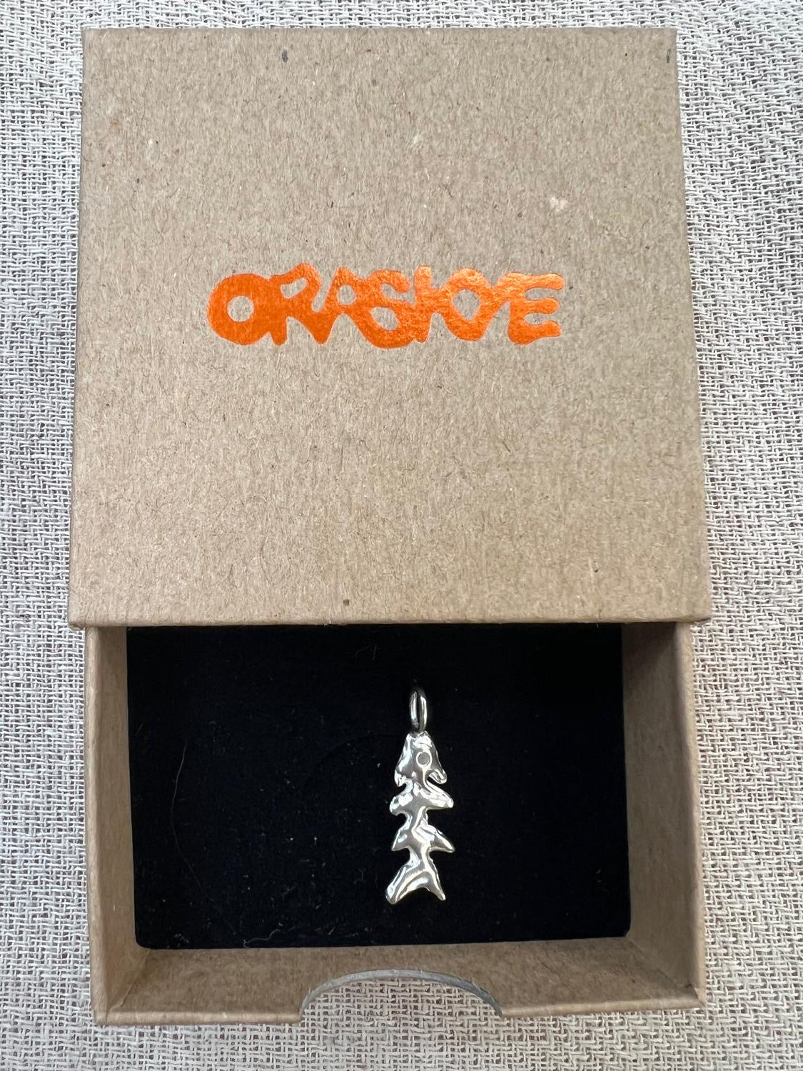 Silver fish pendant in a box with 'ORASKYE' branding on a textured surface